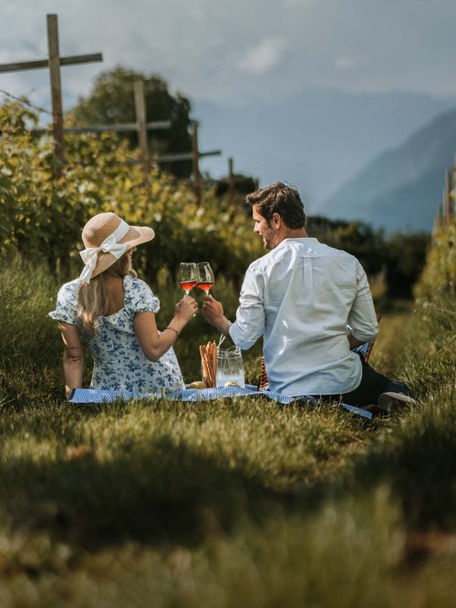 Lingers – Ihr Hotel in Girlan Paar genießt ein Picknick mit Wein in einem Weinberg bei sonnigem Wetter