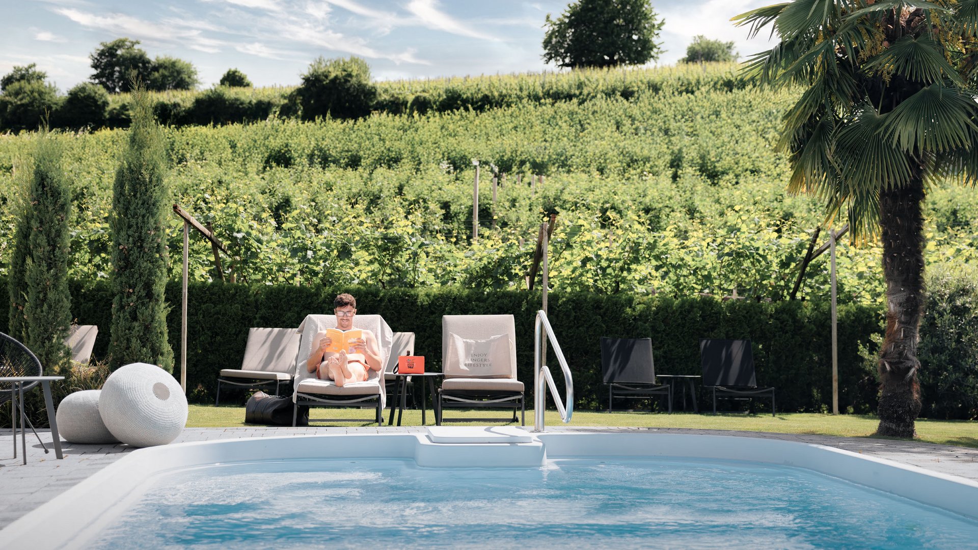 Lingers – Ihr Hotel in Girlan Mann liest am Pool auf einer Liege mit Blick auf grüne Weinberge